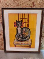 Framed lithograph of a colorful stylized cat sitting on a patterned cushion on a wooden chair with a bright yellow background