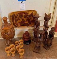 Photo showing carved wood folk art items including six wooden egg cups, a lidded carved container, six statues of people, one small seated figure statue, and a rectangular tray with floral carving.