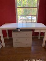 White rectangular desk table with a single drawer on the right side and four legs, situated near a window with natural light.