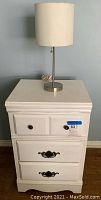 Full front view of white wood nightstand with a desk lamp on top, showing drawer handles and lamp design.