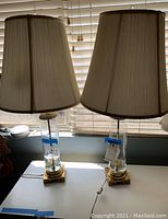 Pair of lamps shown side by side with shades on, placed on a window sill with blinds in background.