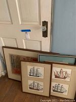 View of several framed sailboat prints and other framed artworks leaning against a door indoors.