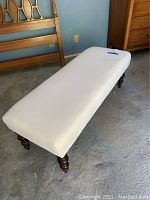 Photo of the white upholstered bench showing the rectangular shape, cushioned top, and dark wooden turned legs.