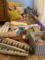 Wide view showing collection of pillows, fabrics, and wallpaper rolls stacked on the floor near a cabinet and window.