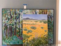 Two framed colorful paintings leaning against a gray wall. One painting shows a forest scene with varied colors and thick brush strokes. The other painting has a yellow flower field with hills and sky in the background.