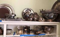 Wide view on two shelves showing assorted tarnished silver, silverplate, pewter trays, pitchers, bowls, teapots and decorative items