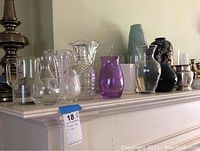 Photo showing various clear glass and crystal vases including a purple glass vase and a black urn with floral decoration on a mantle.