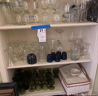 Shelf with mixed clear, green depression, and cobalt blue barware glassware, including glasses, pitchers, and small cups.