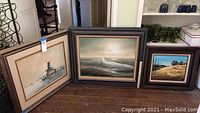Three pieces of framed wall art leaning against a wall, showing a seascape, windmill scene, and rural house painting.