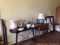 Wide shot showing large vintage floral hand washing pitcher, framed pictures, ceramic pourers, lamp on wooden tables against a wall