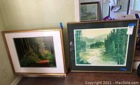 Two framed landscape paintings leaning against a wall, showing nature scenes in green tones.