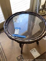 Top view photo of round wood-framed glass table showing carved detail on the wood frame and clear glass top with reflections. Table has some scratches visible on the surface.