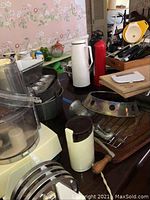 Photo showing several metal pots and pans, a food processor base, plastic kitchen tools, a white thermos, and a red container.