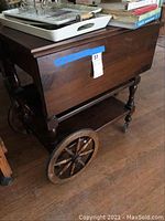 Side view of the vintage wooden bar cart showing spoked wheels and turned legs, with some scratches visible on wood surface. Cart contains some trays and books not included in sale.