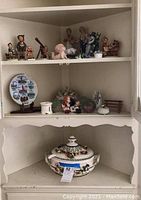 Wide shot of shelves showing all small figurines, soup tureen, souvenir plate, and additional ceramic and glass items.