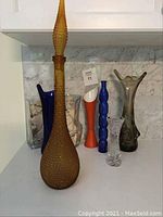 Photo showing six assorted vases including a tall amber hobnail textured vase, a deep blue glass pitcher, and several other colorful vases arranged together.