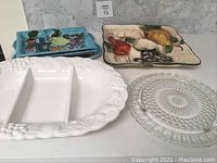 Three platters on a shelf: large hand-painted square ceramic platter with fruit motifs, large white ceramic divided platter, and round clear glass dessert platter.