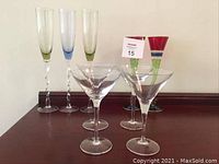 All 9 stemware glasses on a dark wood surface: 4 clear martini glasses in front; behind, 2 colorful flutes and 3 Italian handblown flutes with multicolored stems