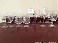Front view of 13 vintage glasses showing 7 etched clear glasses, 2 small violet stemmed glasses, and 4 clear square bowl glasses.