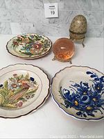 Photo of three hand-painted ceramic wall plates with floral and bird motifs and two decorative table pieces, an egg-shaped gold pattern ornamental and a round pinkish-orange decorative piece.