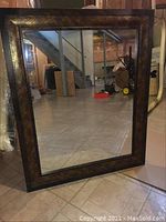 Square brown framed mirror with dark wood style frame, dimensions 28x34 inches, shown standing on a tiled basement floor.