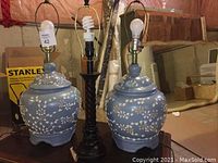 Three table lamps, two grey ceramic with white floral patterns, one black wooden, shown without lampshades. Background includes boxes and other stored items.