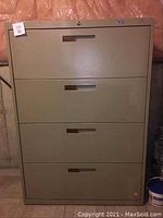 Front view of a khaki tan metal filing cabinet with four drawers, showing recessed handles and label slots. Cabinet is worn with some surface scratches and a small residue spot on top.
