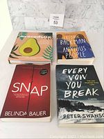 Four assorted books including Avocaderia cookbook and three novels placed on white table against marble background with lot number sign