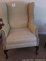 Front view of one beige wing chair showing curved wing back, nailhead trim on arms, light beige fabric upholstery with visible wear.