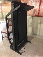 Side view of vintage treadmill folded upright, showing black metal frame and running belt, located in basement environment.