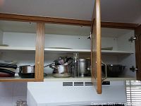 Open kitchen cabinet showing stainless steel cookware including pots, frying pans and glass bowls with lids.