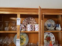 Wide view of wooden shelving unit holding glassware and china items including wine glasses, floral mugs, and plates