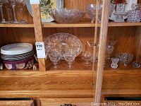 Wooden cabinet shelf with six crystal sherbert cups in front of a large divided serving platter, assorted smaller crystal bowls and bud vases, and plates stacked on the left.