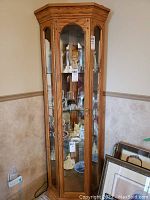 Full view of the oak corner curio cabinet showing angled top, glass front door with key, and visible contents inside on glass shelves.