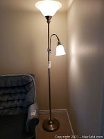 Full view of the floor lamp positioned near a wall and armchair, showing the tall upright light and attached reading light.