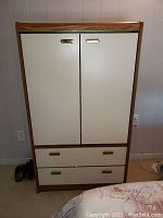 Front view of wardrobe showing wood frame, two white doors, and two bottom drawers with brass recessed handles.