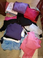 Multiple folded sweaters and tops stacked on a carpeted floor showing various colors like purple, red, pink, black, and white including some light blue fleece material