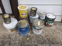 Overall view of paint and stain cans grouped on a concrete floor.