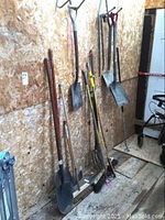 Photo showing an assortment of yard tools leaning against plywood wall including shovels, forks, axe, broom, hoe, pry bars