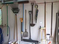 Photo showing various rakes, brooms, shovels, a hoe, a brush, and umbrella hanging on wall hooks.