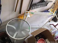Round metal patio side table with frosted glass top and white metal frame seen in cluttered indoor area.