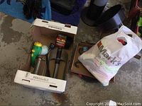 Photo showing a cardboard box containing booster cables with red and black clamps, a garden trowel with a black handle, a large funnel behind the box, and a white bag presumably containing rags. Floor and other storage items visible in background.