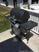 Photo showing black Broil Mate propane BBQ with two burners, closed lid, wheels, and side shelf, outside on concrete patio in sunlight.