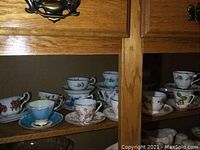 Photo showing multiple teacup and saucer sets on wooden shelves with floral and decorative patterns.