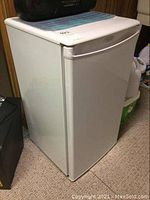 White Danby bar fridge shown from front and side angles, placed on carpeted floor against a wood-paneled wall. Integrated handle and brand logo visible.