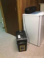 Photo of paper shredder and radio placed on carpet near wooden door and white cabinet. Radio has power cord plugged in and is on.