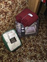 Photo showing burgundy fleece comforter set packaged, white quilted sheet set also packaged, and small afghan throw under packaging on patterned carpet