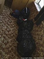 Six large black garbage bags filled with ladies clothing, placed on carpet by a wall and couch.