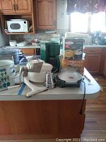Photo of kitchen table with various small appliances and kitchenware, including Salsa Master, Krups coffee maker, grinder, waffle maker, salad spinner, and mixing bowls.
