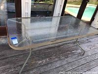 Side angled view of large rectangular glass top patio table with metal frame and legs on a wooden deck showing weathered, used condition.